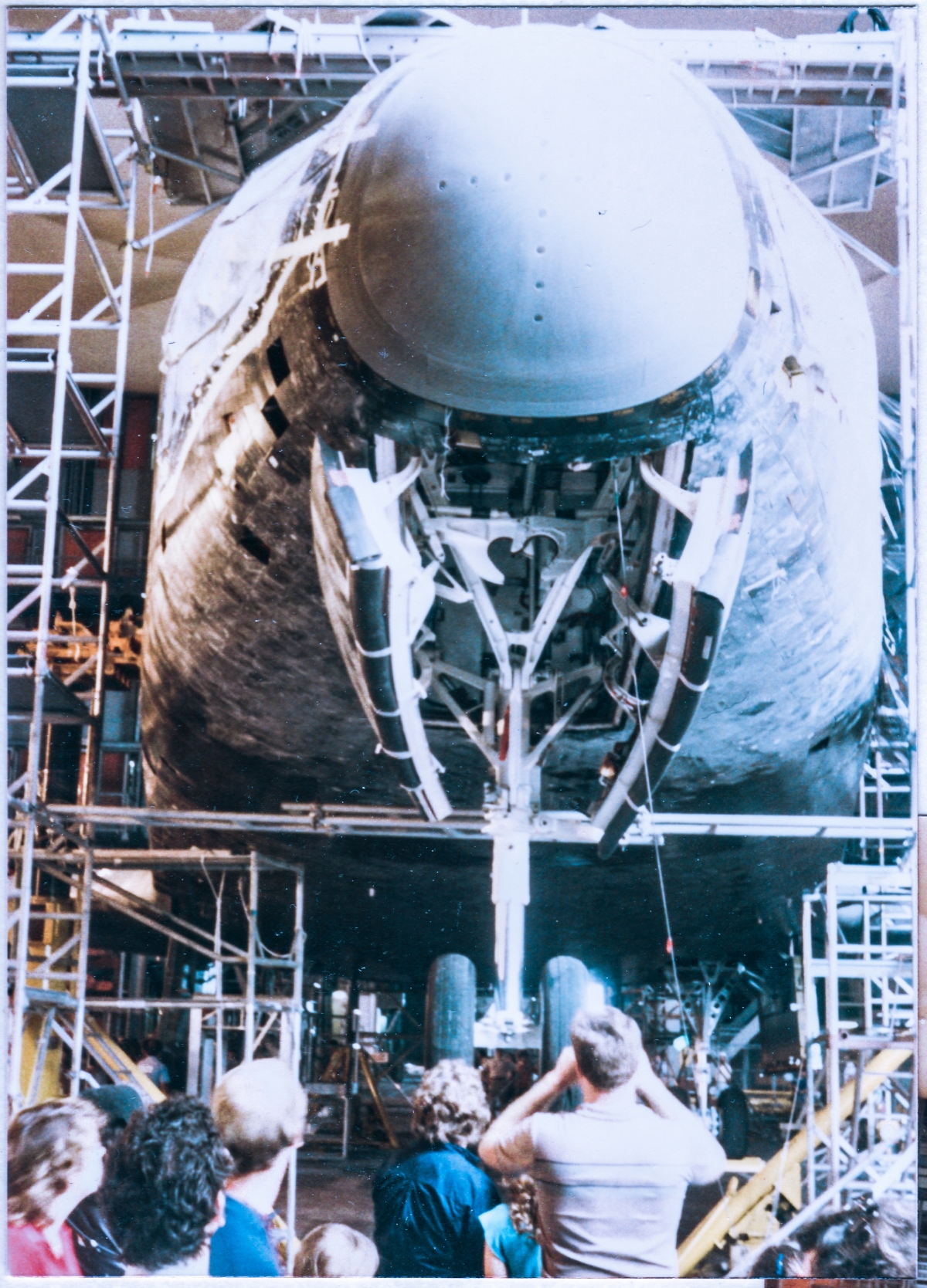 In front and beneath the Nose Cap of the Space Shuttle Columbia, inside the Vehicle Assembly Building at Kennedy Space Center, Florida. Below, the open doors for the nose landing gear reveal some of the hardware associated with that nose gear, including the locking mechanisms. Columbia is not supported by its landing gear, but instead is supported by lifting stanchions (which are visible in the other pictures on this page), which allows servicing of the gear, and much else, without need for further arrangements to enable such servicing by taking the system(s) in question out of any load-bearing configuration. 